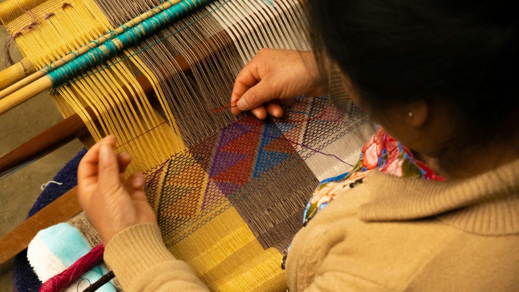 over the shoulder image of a woman intricately weaving a piece of thread through a loom to make a piece of decorative fabric